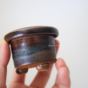 May include: A small, cylindrical ceramic pot with a wide, flat rim and three short, stubby feet. The glaze features a blend of dark brown, earthy tones, and subtle hints of teal. The text "leaves in clay" is printed in white on the side.