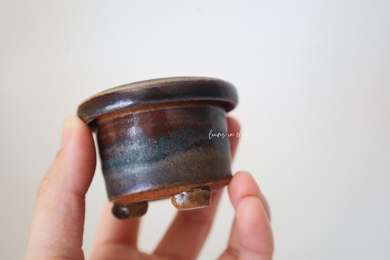 May include: A small, cylindrical ceramic pot with a wide, flat rim and three short, stubby feet. The glaze features a blend of dark brown, earthy tones, and subtle hints of teal. The text "leaves in clay" is printed in white on the side.