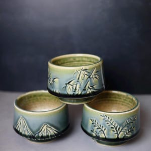 May include: Three ceramic bowls with a green and blue gradient glaze. Each bowl features a distinct embossed design: one with bamboo stalks, one with mountain peaks, and one with fern-like foliage. The bowls have a dark base and a smooth finish.