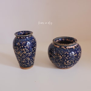 May include: Two ceramic vases with a speckled blue glaze and a light brown rim. One is tall and slender, the other is round and squat. The vases are likely for decorative purposes.