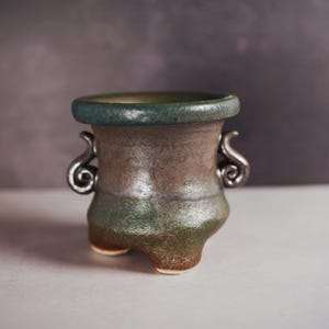 May include: A small, handcrafted ceramic pot with a unique shape and color gradient. The pot features a dark teal rim, a brown and green body, and decorative silver-colored handles. The pot has three small feet.