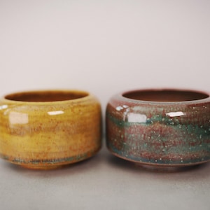 May include: Two small, round ceramic bowls with a smooth, glossy finish. The bowl on the left is a vibrant mustard yellow with subtle brown undertones. The bowl on the right features a blend of deep red, teal, and brown hues with white speckles.
