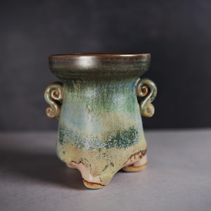 May include: A handcrafted ceramic vessel with a unique, three-legged base. The pottery features a blend of green, blue, and tan hues, with decorative swirl handles. The rim is a darker brown, adding contrast to the piece.
