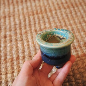 May include: A small, round ceramic pot featuring a gradient glaze that transitions from a deep navy blue at the base to a lighter teal and speckled cream at the wide, flat rim. The vessel is supported by three small, rounded feet at the bottom.