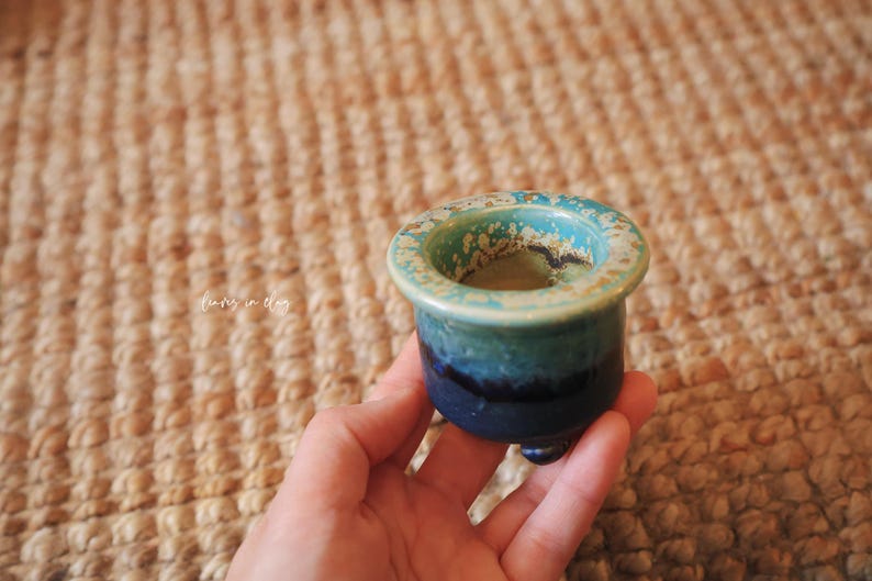 May include: A small, round ceramic pot featuring a gradient glaze that transitions from a deep navy blue at the base to a lighter teal and speckled cream at the wide, flat rim. The vessel is supported by three small, rounded feet at the bottom.