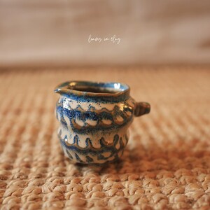 May include: A small, handmade ceramic creamer with a blue and white wave pattern. The creamer has a small handle and a spout for easy pouring. The piece is on a textured, neutral-toned surface.