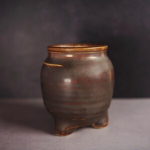 May include: A handmade ceramic pot with a dark brown glaze. The pot has a rounded body, a slightly flared rim, and three small feet. The pot is approximately 5 inches tall and 4 inches wide.