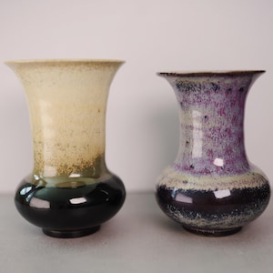 May include: Two ceramic vases with flared rims and rounded bodies. The left vase features a cream-colored top transitioning into a dark green base. The right vase displays a mottled purple and white upper section with a dark, speckled base.