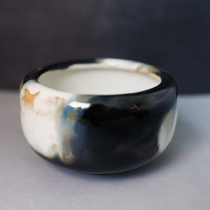 May include: A small, round ceramic bowl with a smooth, glossy finish. The exterior features a marbled pattern of deep black, creamy white, and subtle hints of golden-brown. The interior of the bowl is a solid, clean white with a curved rim.