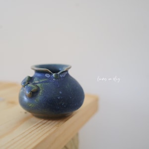 May include: A small, rounded ceramic pitcher with a deep blue glaze and mottled green accents. The vessel features a distinct pouring spout and a small, sculpted leaf-like detail on the side. The text "leaves in clay" is printed in white.