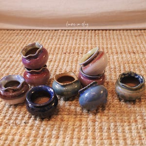 May include: A collection of small, handcrafted ceramic pots in various colors, including blue, purple, and tan. The pots have a unique, wavy rim design and are arranged on a textured surface.