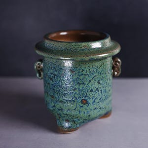 May include: A small, ceramic pot with a textured, speckled green and blue glaze. The pot has a wide rim, two small handles, and three feet. The interior is a warm brown color.