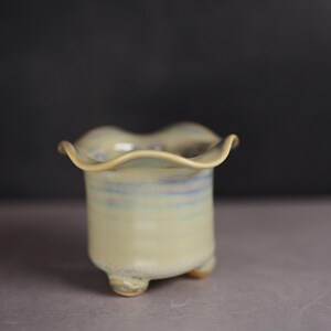 May include: A small ceramic planter with a ruffled, wavy rim and a pale yellow glaze featuring subtle blue and purple undertones. The vessel has a cylindrical body supported by three small, rounded feet at the base, creating a stable structure.