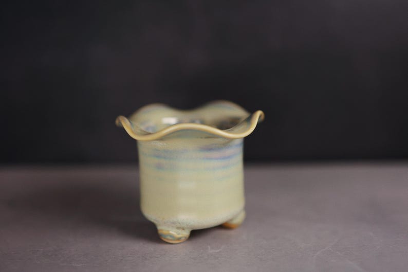 May include: A small ceramic planter with a ruffled, wavy rim and a pale yellow glaze featuring subtle blue and purple undertones. The vessel has a cylindrical body supported by three small, rounded feet at the base, creating a stable structure.