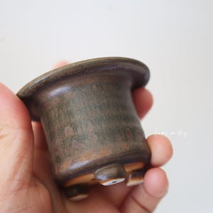 May include: A small, cylindrical ceramic pot with a wide, flared rim and a dark, mottled brown and green glaze. The vessel features three short, rounded feet at the base, each with a small, circular white accent on the underside.