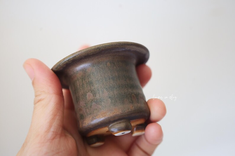 May include: A small, cylindrical ceramic pot with a wide, flared rim and a dark, mottled brown and green glaze. The vessel features three short, rounded feet at the base, each with a small, circular white accent on the underside.
