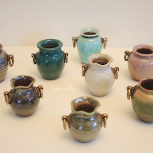 May include: A collection of eight small ceramic vases in various colors, including blue, green, and pink. Each vase has a unique shape and is adorned with bronze-colored handles. The vases are arranged on a white surface.