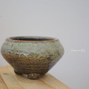 May include: A handcrafted ceramic bowl with a unique glaze. The bowl features a rounded shape with a wide opening and sits on three small feet. The exterior has a mottled green and brown finish, giving it a rustic appearance. The bowl is suitable for plants or decorative use.