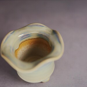 May include: A small ceramic vessel with a wavy, flared rim. The exterior features a pale, off-white glaze with subtle blue accents. The interior transitions from a light blue rim to a warm, golden-yellow base, creating a smooth color gradient.