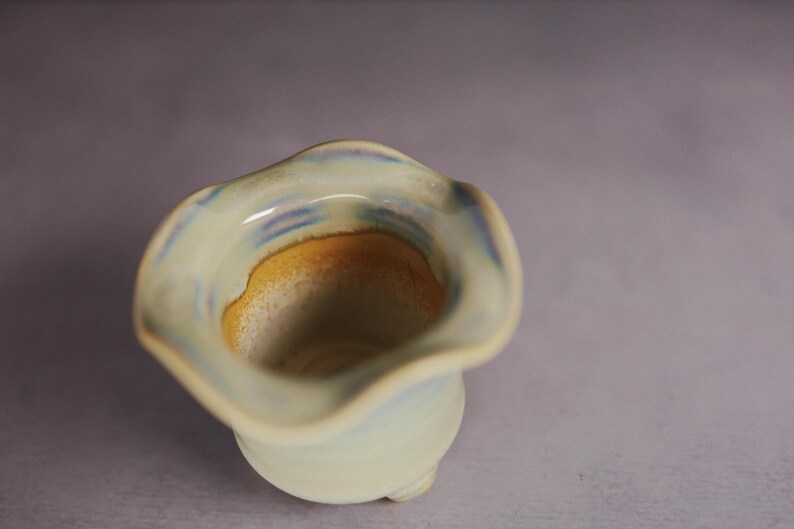 May include: A small ceramic vessel with a wavy, flared rim. The exterior features a pale, off-white glaze with subtle blue accents. The interior transitions from a light blue rim to a warm, golden-yellow base, creating a smooth color gradient.