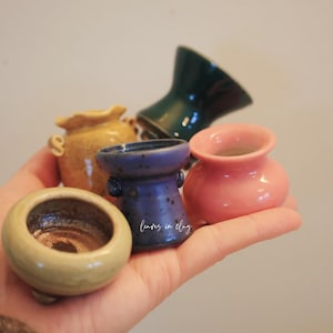 May include: A collection of small, colorful ceramic vases in various shapes and sizes. The vases are in shades of yellow, blue, pink, and green. The vases are held in a hand.