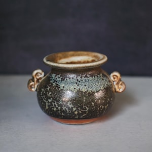 May include: A small, handcrafted ceramic pot with a rounded body and two ornate handles. The pot features a speckled, dark brown glaze with hints of blue and a lighter rim. The base is a reddish-brown color.