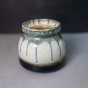 May include: A small ceramic pot with a rounded body and a wide opening. The glaze features a white base with blue-grey drips running down the sides, transitioning into a dark brown and black band at the base and a textured rim at the top.