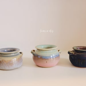 May include: Three ceramic pots in a row, each with a unique color scheme. The pots have small handles and a rounded shape. The colors include blue, pink, and dark blue, with a light background.