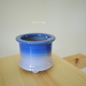 May include: A small ceramic pot with a smooth, glossy finish that transitions from a deep blue at the rim to a pale, light blue at the base. The pot features a wide, rounded rim and a small, raised foot. The text "leaves in clay" is visible.