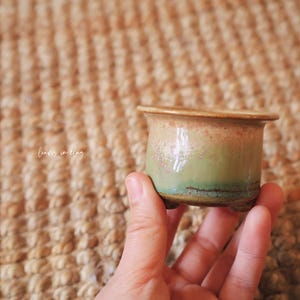 May include: A small ceramic pot with a flared rim and three short feet. The glaze features a gradient transition from a soft peach-pink at the top to a muted sage green at the base, with a dark brown horizontal line accenting the lower section.