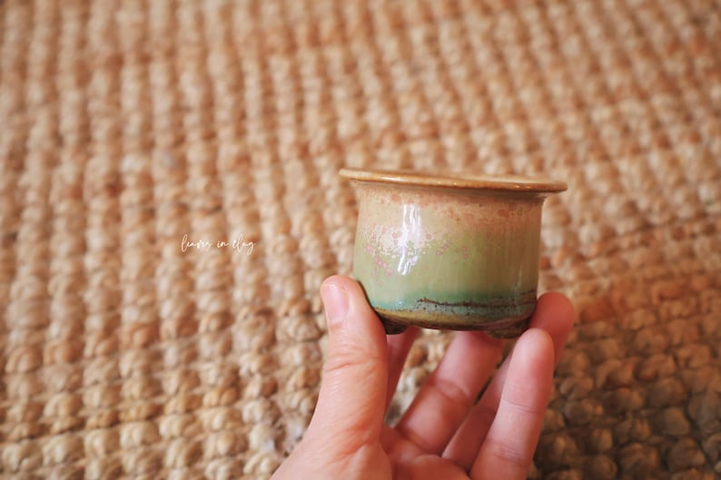 May include: A small ceramic pot with a flared rim and three short feet. The glaze features a gradient transition from a soft peach-pink at the top to a muted sage green at the base, with a dark brown horizontal line accenting the lower section.