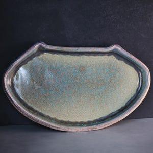May include: A ceramic serving platter with a unique shape and a speckled teal glaze. The rim is a darker shade of teal with a silver-colored edge. The platter is approximately 12 inches long and 8 inches wide.