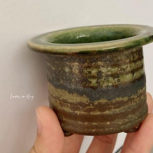 May include: A small, cylindrical ceramic planter with a wide, flat rim and three short feet. The exterior features a textured, mottled glaze in shades of earthy brown, dark gray, and moss green. The text "leaves in clay" is printed on the side.