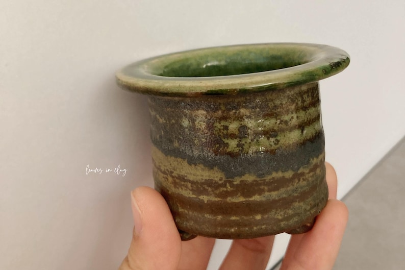 May include: A small, cylindrical ceramic planter with a wide, flat rim and three short feet. The exterior features a textured, mottled glaze in shades of earthy brown, dark gray, and moss green. The text "leaves in clay" is printed on the side.