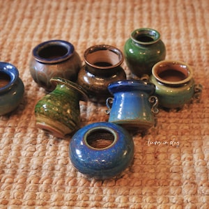 May include: A collection of small, handcrafted ceramic pots in various colors, including blue, green, and brown. The pots have a rustic, textured appearance and are arranged on a woven surface. These decorative pots are ideal for small plants.