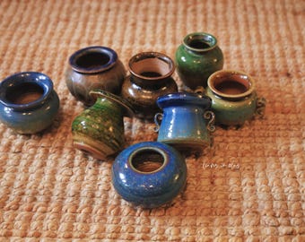 Puede incluir: Conjunto de pequeñas macetas de cerámica hechas a mano en varios colores, incluyendo azul, verde y marrón. Las macetas tienen un aspecto rústico y texturizado, dispuestas sobre una superficie tejida. Ideales para plantas pequeñas.