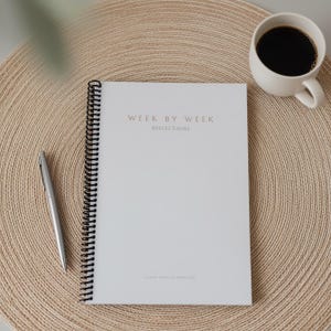 May include: A white spiral-bound notebook with gold lettering reading "WEEK BY WEEK REFLECTIONS". A silver pen lies next to the notebook on a beige woven mat. A white mug filled with black coffee is in the top right corner.