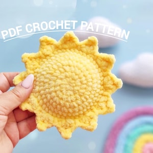 May include: A yellow crochet sun with a textured surface is held in a hand. The sun has a scalloped edge and is set against a light blue background with the text "PDF CROCHET PATTERN". A rainbow and clouds are also visible.