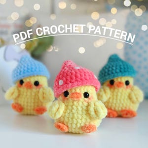 Könnte beinhalten: Drei gelbe Häkelküken mit bunten Hüten. Die Küken haben orangefarbene Schnäbel und Füße sowie schwarze Augen. Die Hüte sind blau, rosa und türkis. Der Text "PDF CROCHET PATTERN" ist über den Küken angeordnet.