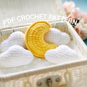 May include: A white wicker basket contains a yellow crocheted crescent moon and four white crocheted cloud shapes. The text "PDF CROCHET PATTERN" is displayed across the top of the image. Soft pink flowers are visible in the background.