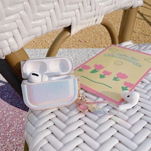 May include: A white AirPod case with an open lid and a shimmering, iridescent cover rests on a white woven chair. A pair of white AirPods and a card with pink flowers and text are also visible.
