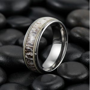 Könnte beinhalten: Nahaufnahme eines silbernen Rings mit einem Band aus hellbraunem und weißem Material. Der Ring hat einen dünnen, goldfarbenen Rand und ist vor einem Hintergrund aus schwarzen Steinen platziert. Der Ring ist ein Schmuckstück.