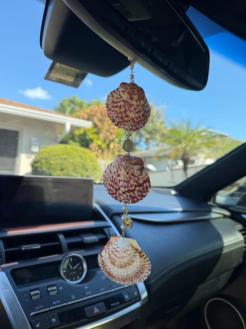 May include: A hanging car ornament featuring three natural scallop shells with reddish-brown patterns, connected by a delicate gold-toned chain with small white beads and circular metal accents, suspended from a vehicle rearview mirror.
