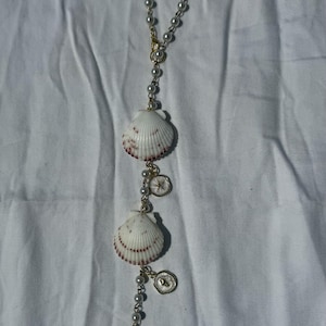 May include: A hanging ornament featuring a gold-toned chain strung with white faux pearls, three white scallop shells with reddish-brown markings, and two small circular gold-toned charms with embossed star and heart details.