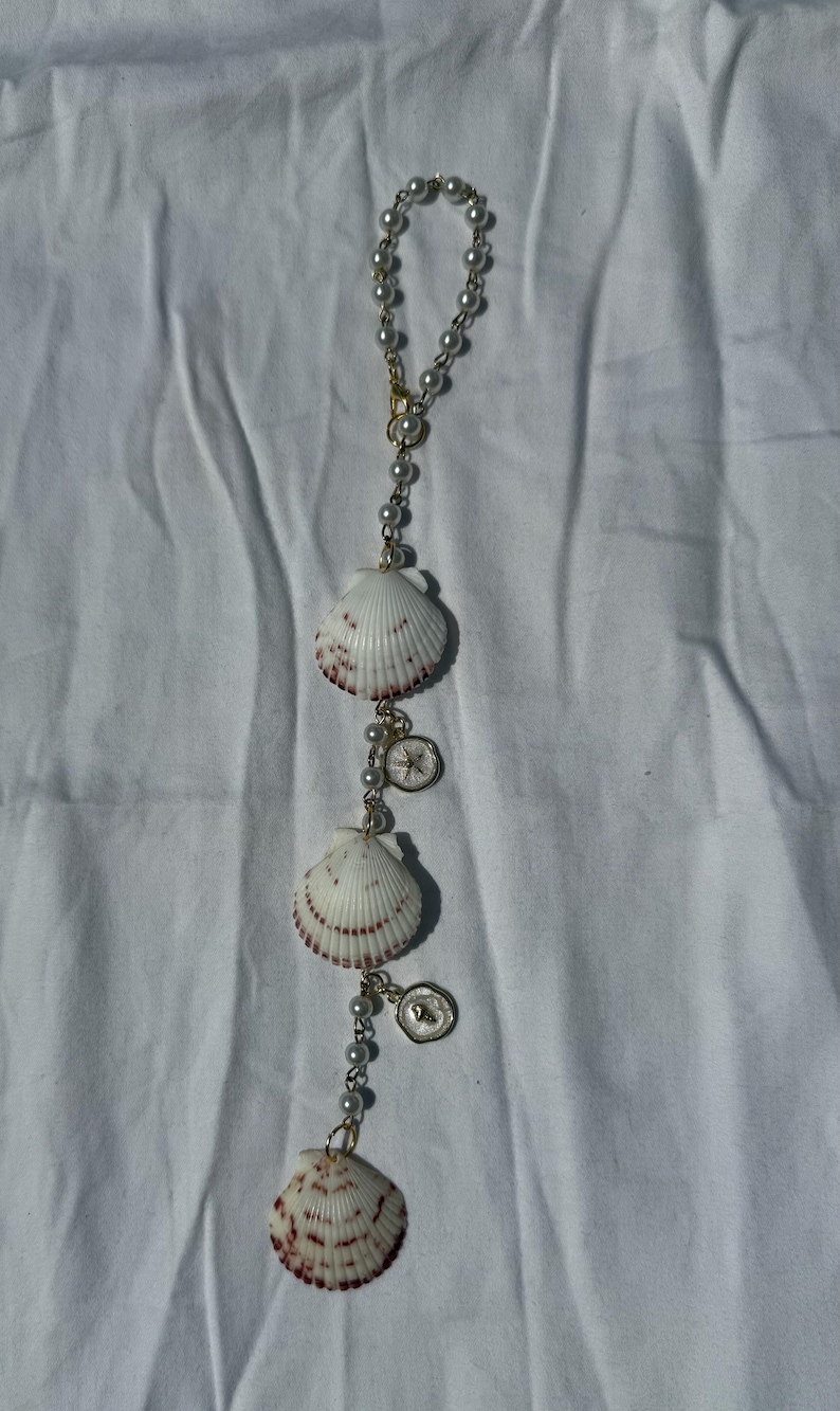 May include: A hanging ornament featuring a gold-toned chain strung with white faux pearls, three white scallop shells with reddish-brown markings, and two small circular gold-toned charms with embossed star and heart details.