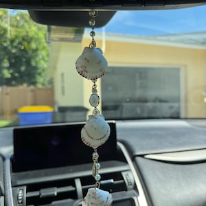 May include: A hanging car ornament featuring three white scallop shells with reddish-brown markings, interspersed with small white faux pearls and a circular gold-toned charm with a star emblem, all connected by a thin gold-toned metal chain.