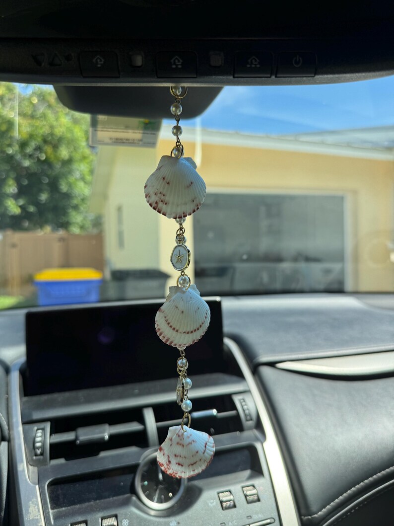 May include: A hanging car ornament featuring three white scallop shells with reddish-brown markings, interspersed with small white faux pearls and a circular gold-toned charm with a star emblem, all connected by a thin gold-toned metal chain.