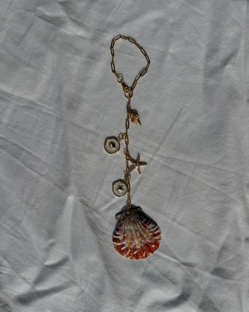 May include: A gold-toned chain charm featuring a series of nautical-themed pendants, including a small shell, a starfish, two circular white enamel charms with gold centers, and a large, textured, fan-shaped seashell with orange and white stripes.