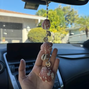May include: A hanging car ornament featuring three fan-shaped, reddish-brown and white striped seashells interspersed with smaller white cowrie shells and small, round, cream-colored beads strung together on a thin gold-toned metal chain.