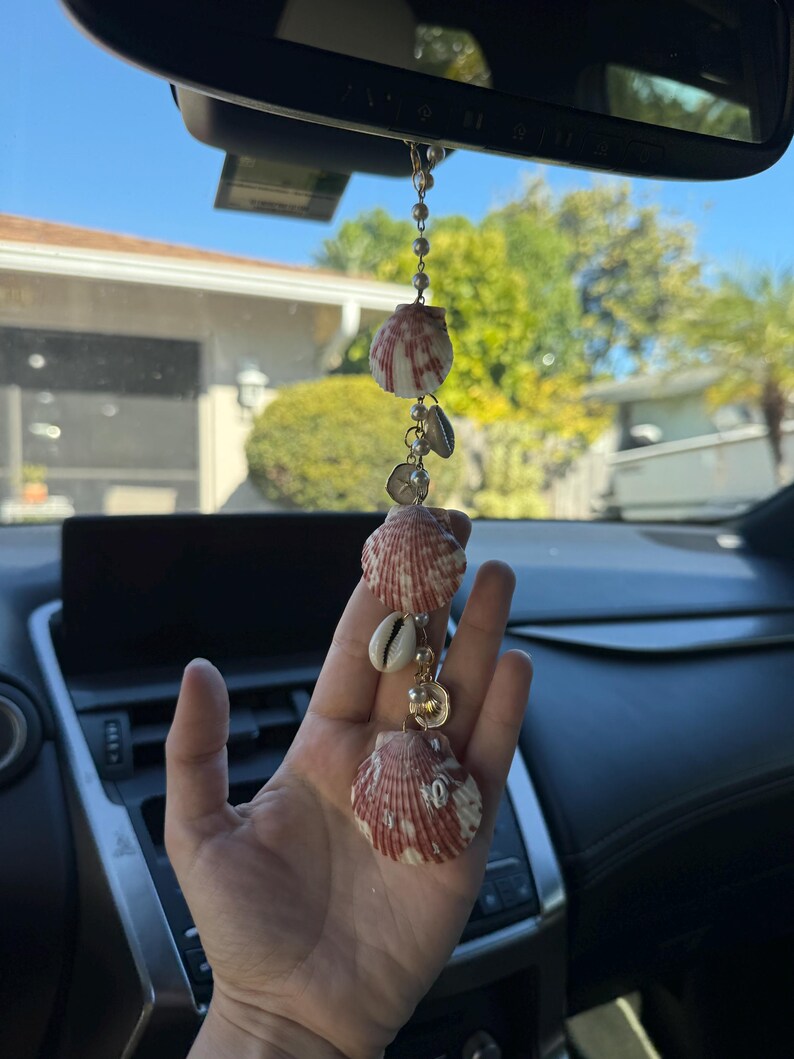 May include: A hanging car ornament featuring three fan-shaped, reddish-brown and white striped seashells interspersed with smaller white cowrie shells and small, round, cream-colored beads strung together on a thin gold-toned metal chain.